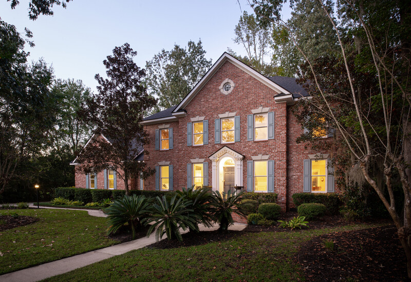 Two-story brick house with illuminated windows photographed at dusk surrounded by mature trees and landscaping.