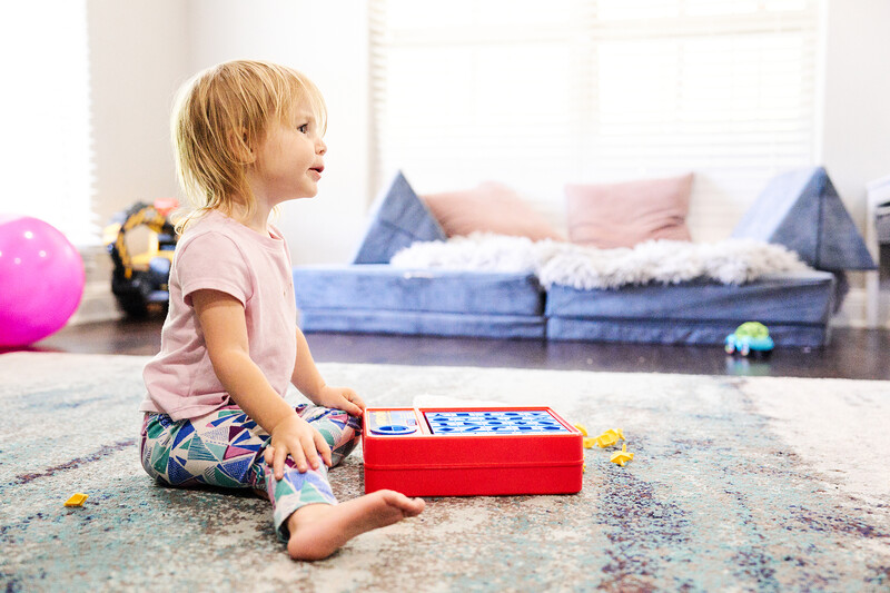 Toddler sitting on carpet playing with a red toy box in a bright playroom.