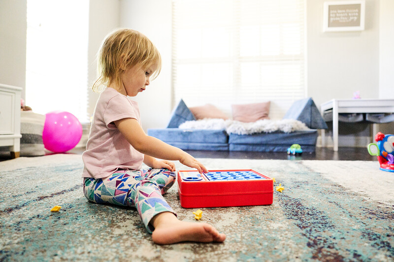 Toddler sitting on a patterned carpet playing with a red toy box in a bright living room.