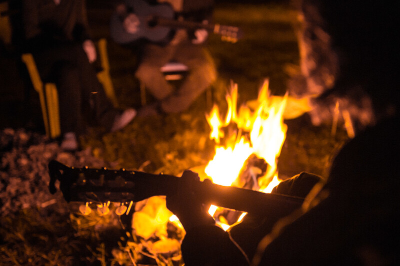 Campfire with bright yellow and orange flames consuming logs at night, with blurred figures visible in background.