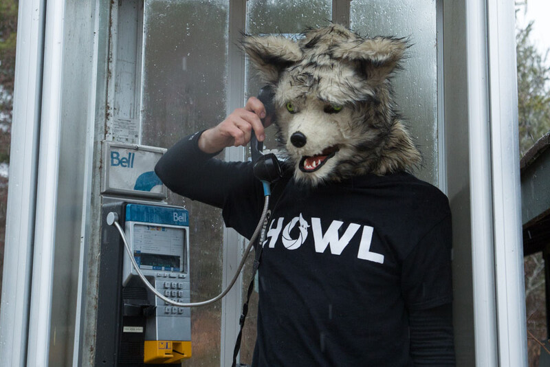 Person in wolf mask and black shirt holding telephone receiver at public phone booth.
