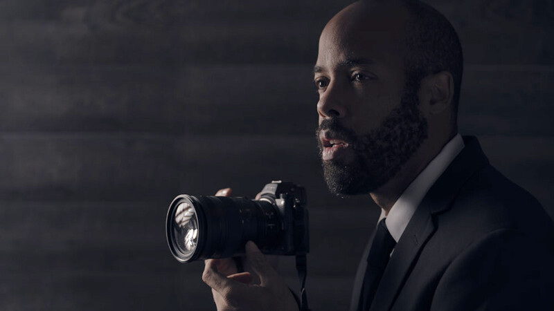 Professional photographer in dark suit holding DSLR camera with mounted flash against dark background.