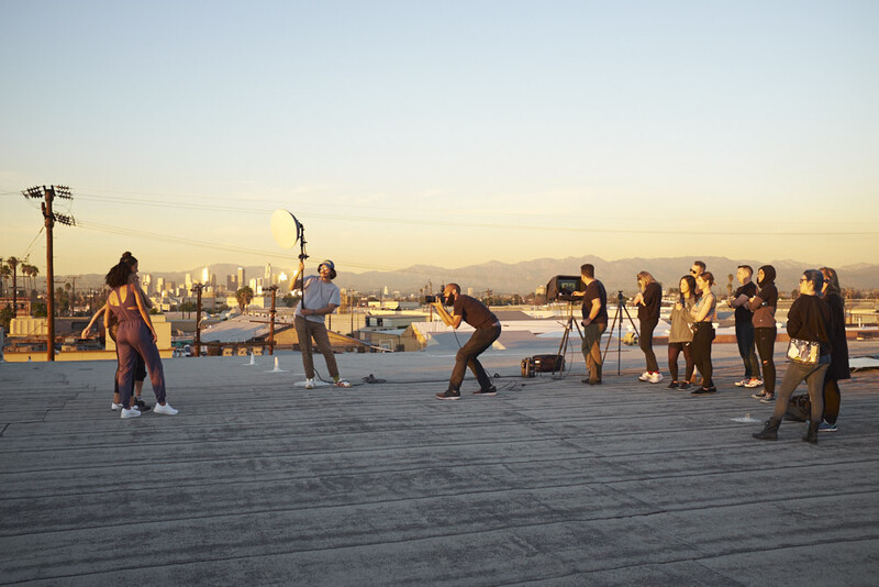 Film crew setting up equipment on a rooftop at golden hour with city skyline and mountains in the background.