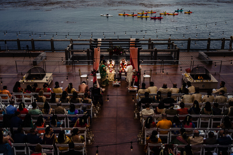 Aerial view of a waterfront ceremony with guests seated in rows facing an altar on a wooden pier.