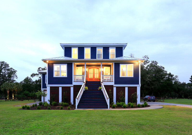 Modern elevated house with dark blue exterior, white trim, and illuminated orange front door at dusk.