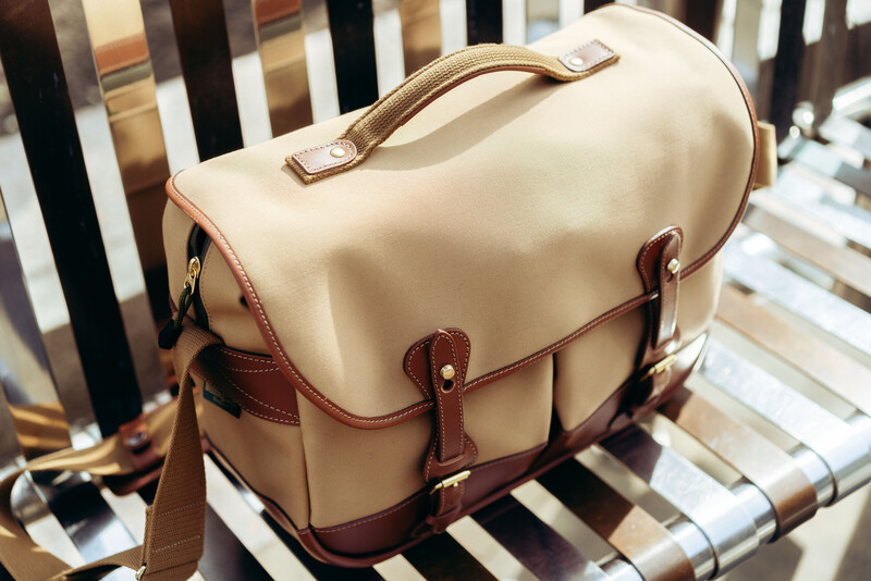 Cream and brown leather camera bag with dual handles and brass hardware on striped background.