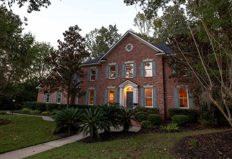 Two-story brick colonial house photographed at dusk with warm interior lighting visible through windows.
