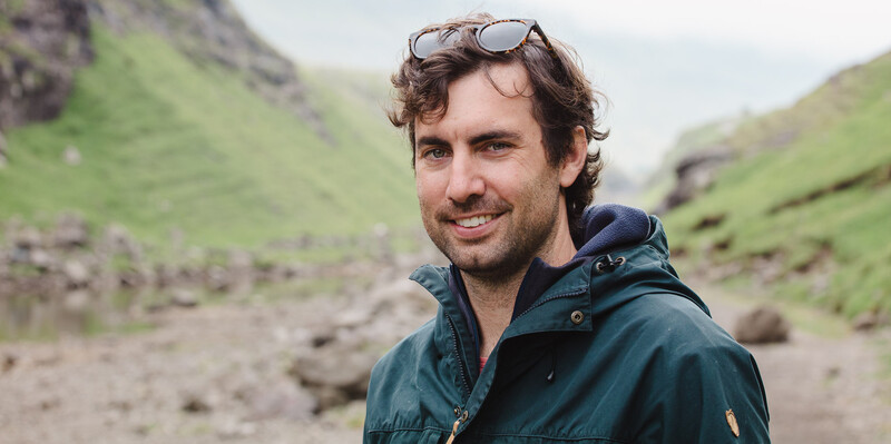 Man smiling at camera in mountainous valley with green slopes and rocky terrain.