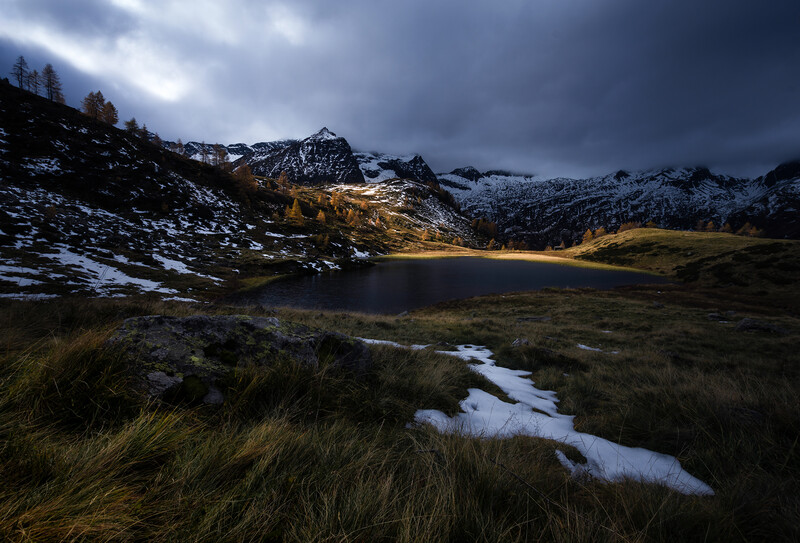 Alpine lake reflecting golden larch trees beneath snow-capped peaks under dramatic storm clouds.