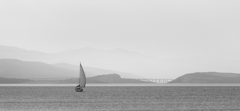 Sailboat with raised sail on calm water, distant bridge and hillsides visible in monochromatic landscape.