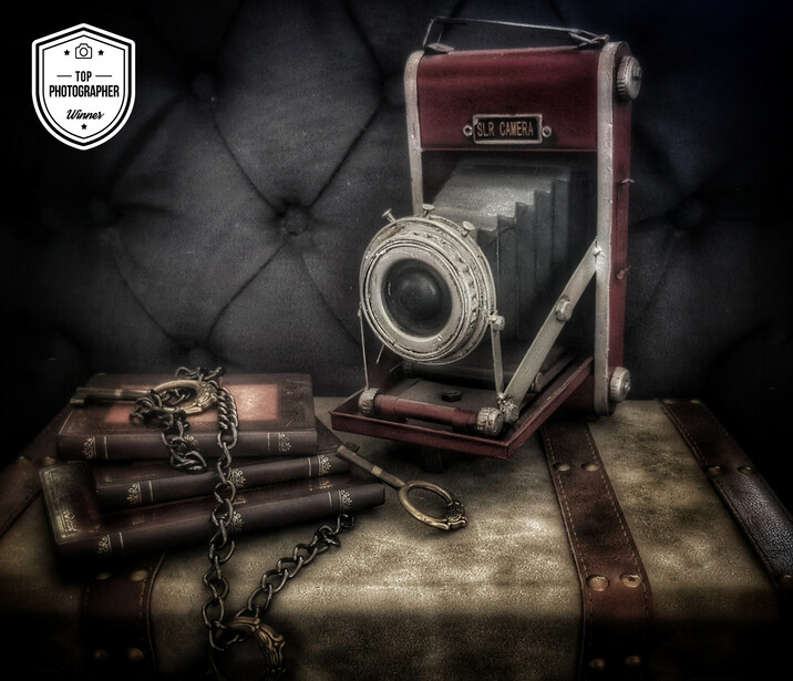 Vintage folding camera on weathered luggage with chain and old books arranged in moody still life.