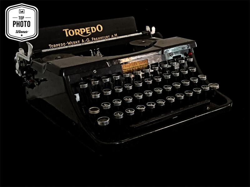 Vintage Torpedo typewriter with black body and round keys photographed against black background.