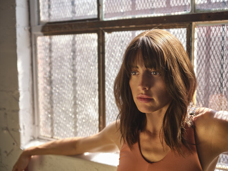 Woman with shoulder-length brunette hair and bangs sitting by a window with wire-mesh panes.