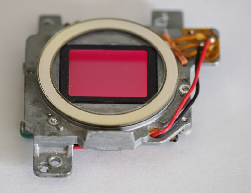 Disassembled camera image sensor with pink filter mounted in metal housing and gold contacts.