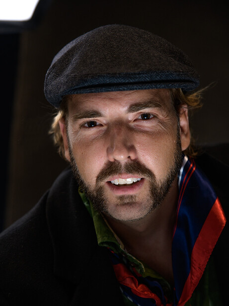 Portrait of a bearded man wearing a flat cap and colorful striped jacket against dark background.