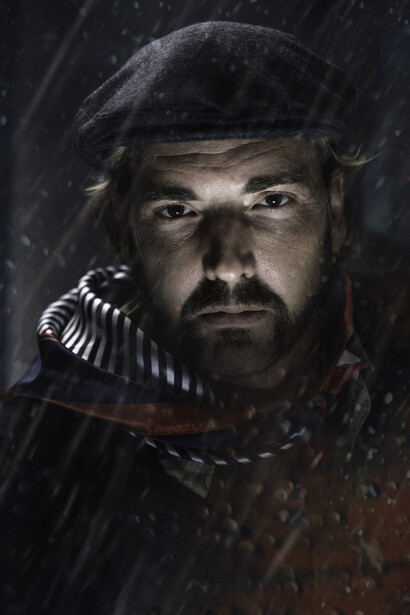 Portrait of a bearded man in period military attire with ornate shoulder epaulettes, photographed in rain with dramatic side lighting.