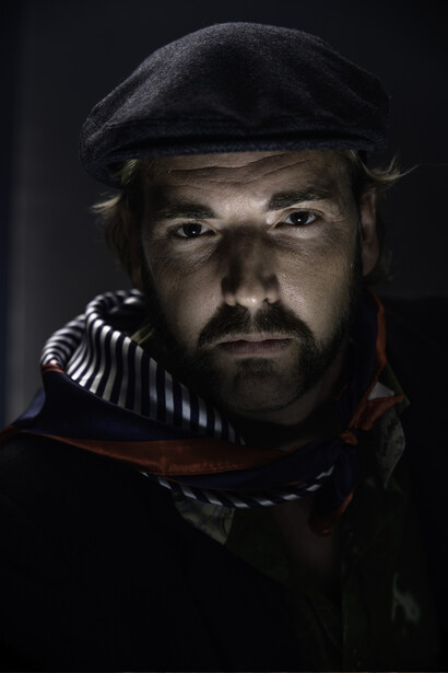 Bearded man in flat cap and period costume against dark background.