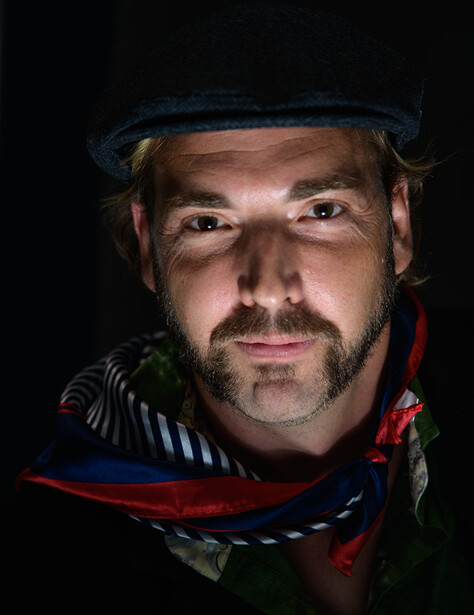 Portrait of a man wearing a dark cap and red climbing harness against a black background.