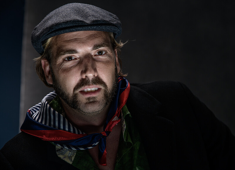 Man wearing a flat cap and dark jacket with red collar against dark background.