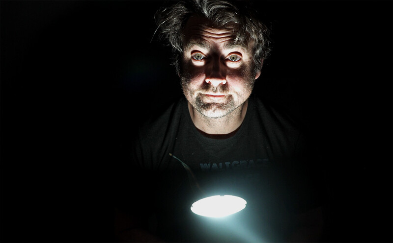 Man's face illuminated by bright LED light source held at chest level against black background.