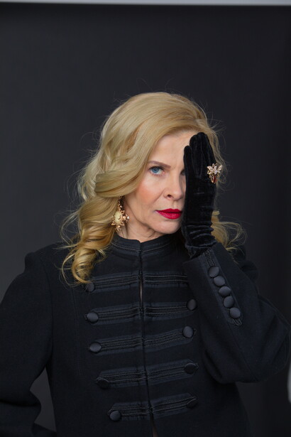 Woman in black military-style jacket against dark background, blonde wavy hair and red lipstick.