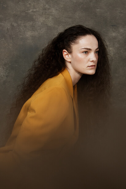 Woman with long dark hair wearing mustard-yellow garment, photographed in profile against neutral textured background.