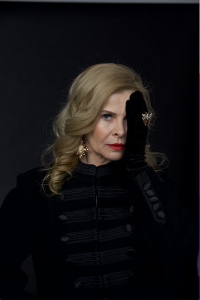 Woman with blonde wavy hair wearing a black quilted jacket against a dark background.