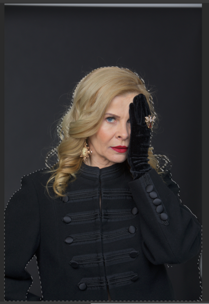 Woman with blonde wavy hair wearing black military-style jacket against dark backdrop.