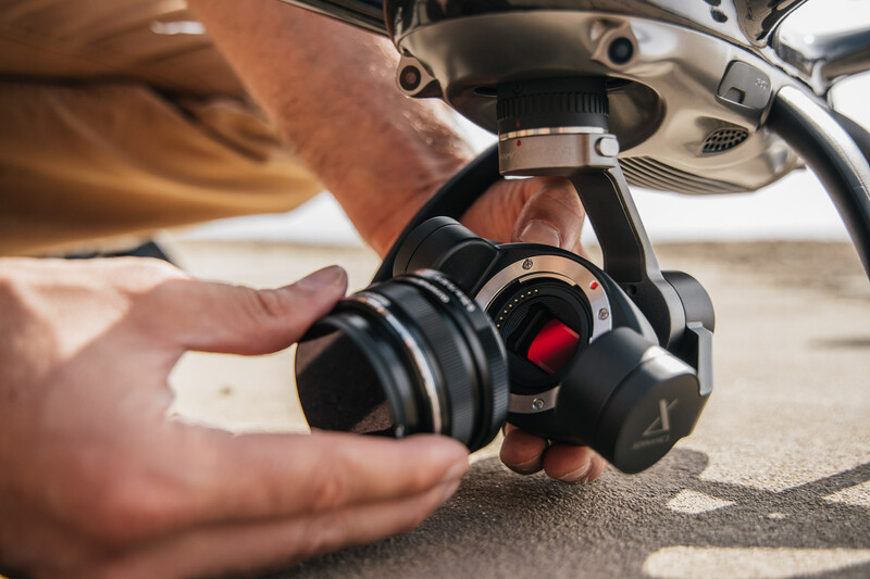 Hands holding a gimbal-mounted camera with red accent details beneath a drone.