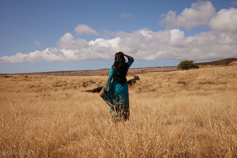 Person in turquoise jacket standing in dry grassland, shielding eyes while looking toward horizon.