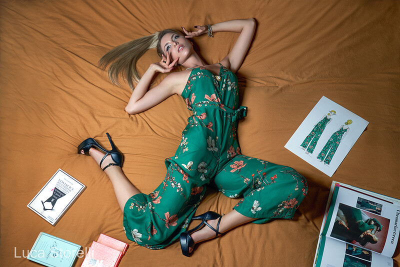 Woman in green floral jumpsuit lying on tan fabric with fashion sketches and mood boards arranged around her.