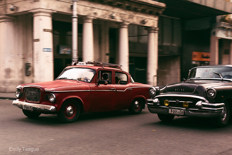 Two vintage automobiles parked on a city street with classical architecture in the background.
