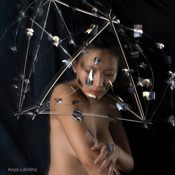 Shirtless person with face paint surrounded by geometric wireframe structure and hanging sculptural elements against black background.