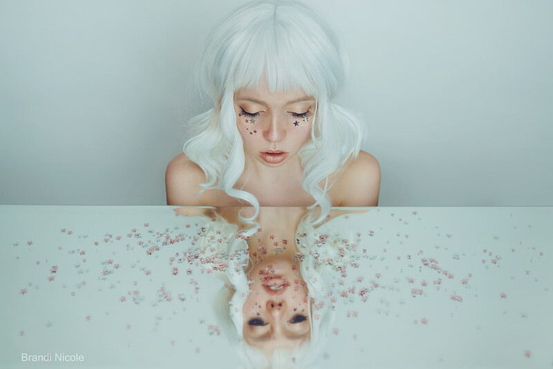 Woman with white wavy hair and glitter makeup looking down at her mirrored reflection on a white surface scattered with pink sparkles.