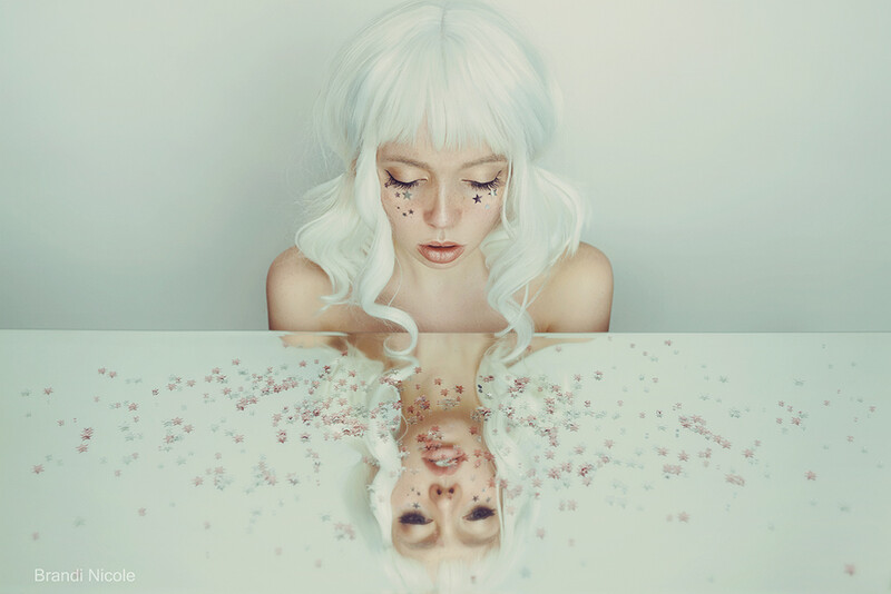 Woman with white hair and glitter makeup looking down at mirror reflection showing scattered sparkles.