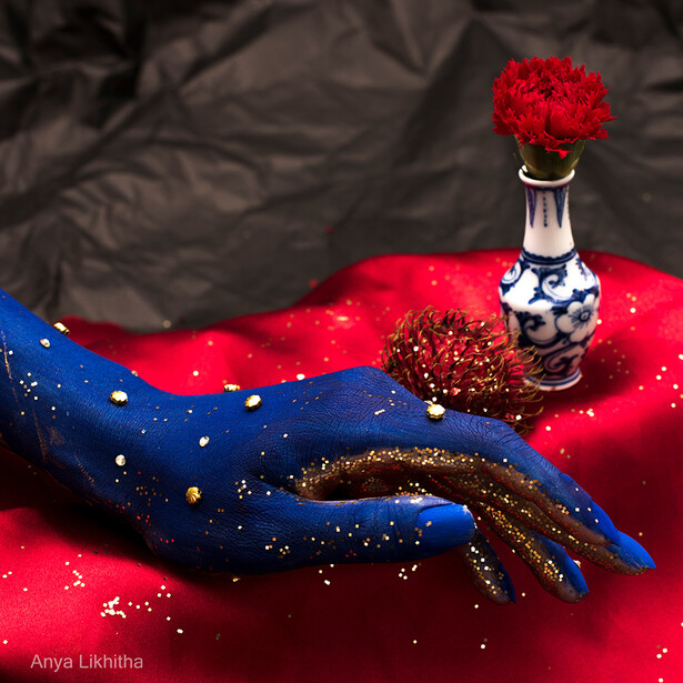 Hand with blue nail art and gold glitter against red fabric with a red flower in a decorative bottle.