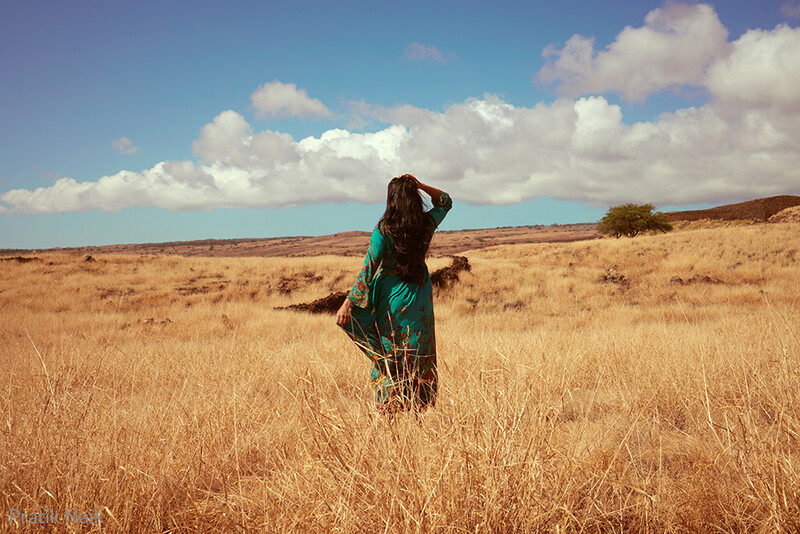 Woman in teal jacket standing in vast golden grassland under blue sky with white clouds.