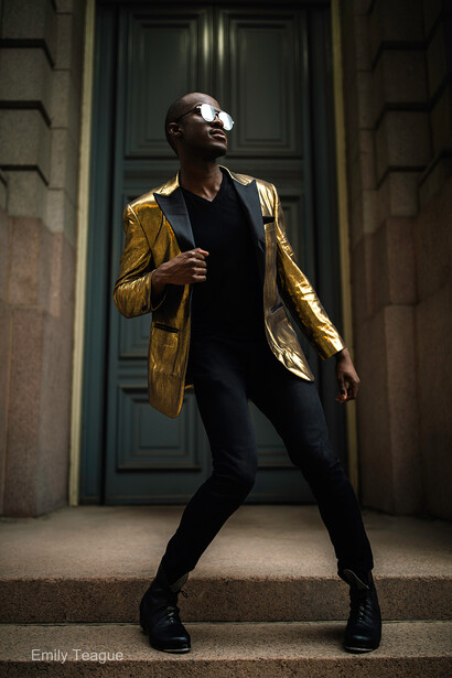 Person in gold jacket and black pants posing against blue-green door in architectural setting.