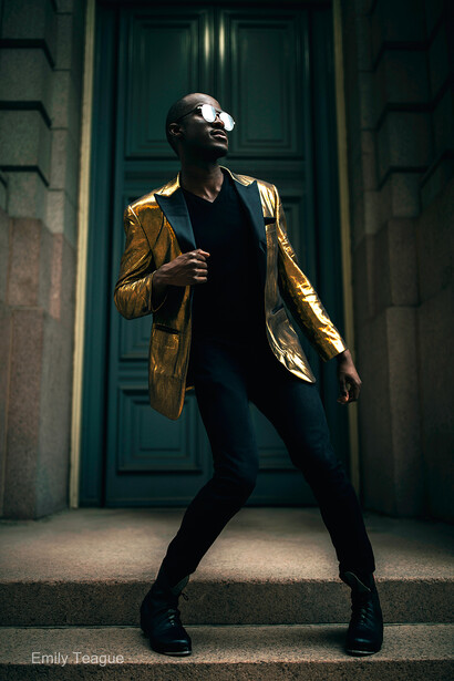 Person in gold metallic blazer and black clothing posed against green doorway.
