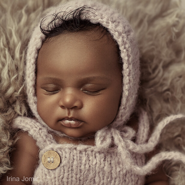 Newborn baby wearing a cream knit bonnet and matching sweater, peacefully sleeping against a textured beige fabric backdrop.