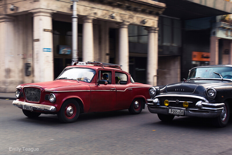 Two vintage cars parked on a city street with colonial architecture in the background.