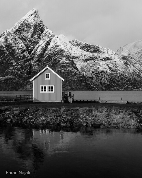 Small white house with dark roof beside still water, with snow-capped mountain rising dramatically behind it.