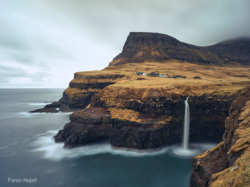 Waterfall cascading from a cliff edge into turquoise water below a moss-covered mountain.