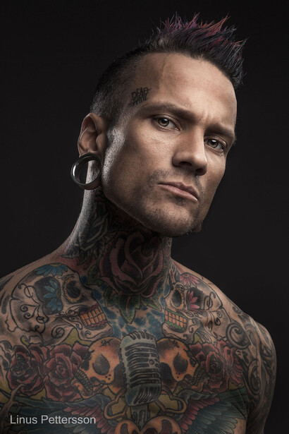 Portrait of a man with extensive tattoo coverage and styled hair against a dark background.