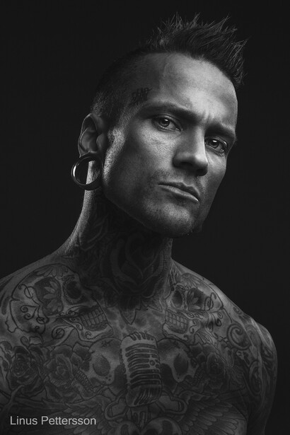 Black and white portrait of a man with styled hair and facial hair, wearing a dark textured shirt with visible tattoos.