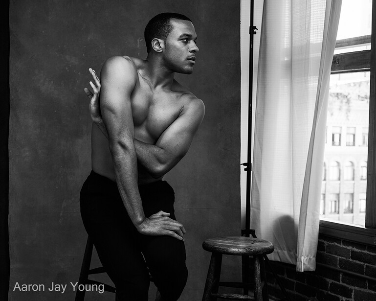 Shirtless man in black pants seated on a stool near a window in a studio setting.