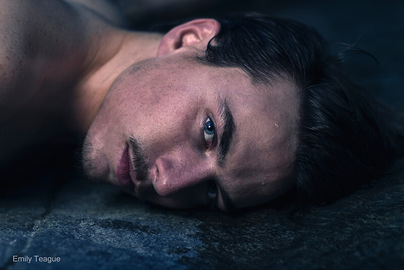 Man lying on dark ground with head tilted back, dramatic moody lighting.