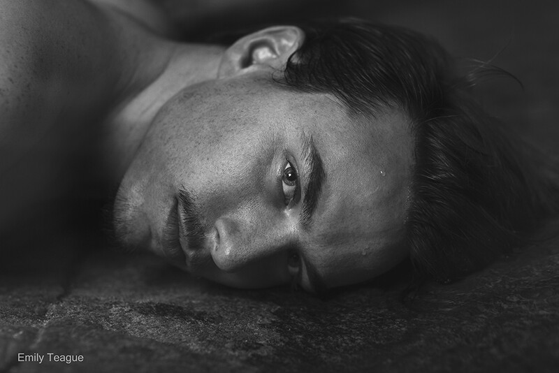 Black and white portrait of a person's face lying on textured ground, eyes gazing upward with dramatic lighting.