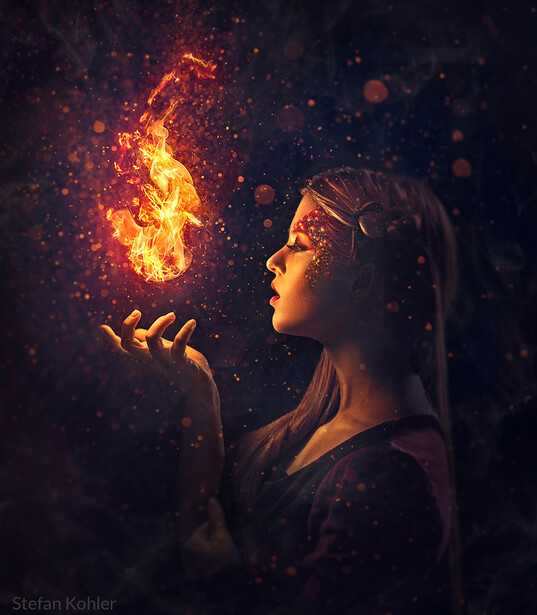 Profile of person with long hair holding glowing fire particles in their palm against dark background.