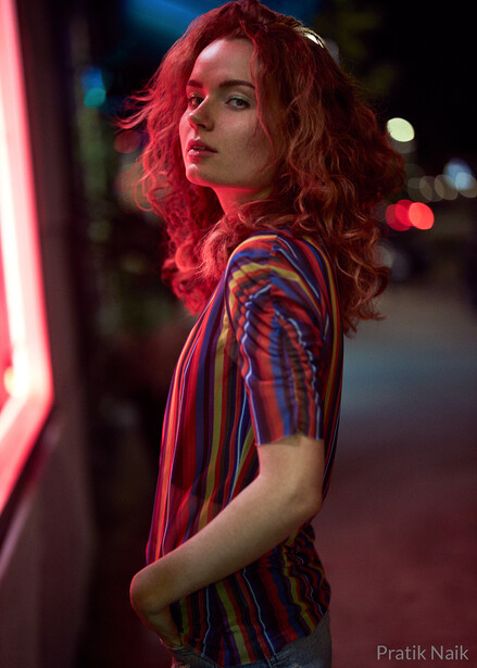 Woman with red curly hair wearing striped shirt, backlit by neon red light in dark urban setting.